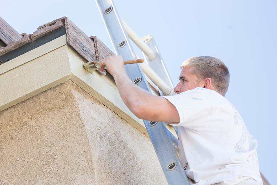 Man on a ladder painting house trim and finishings outside